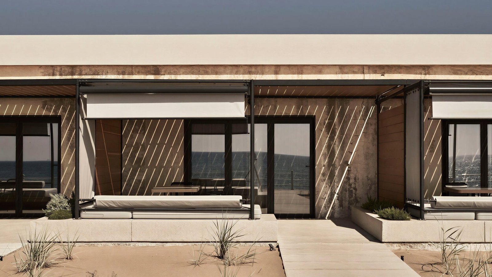 Beachfront WineTank Suite at Dexamenes Seaside Hotel with large glass doors and striped shadows. Sandy yard with sparse grass under a clear blue sky, calm atmosphere.