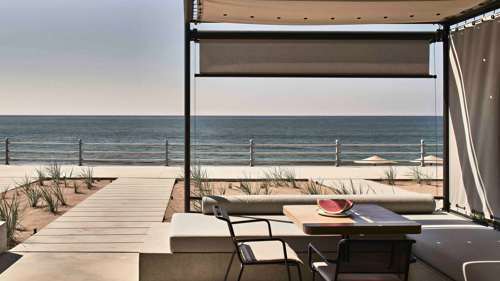 Beachfront WineTank Suite view at Dexamenes Seaside Hotel from a shaded terrace with a table and chairs, a watermelon slice, and a boardwalk leading to the sea under a clear sky.