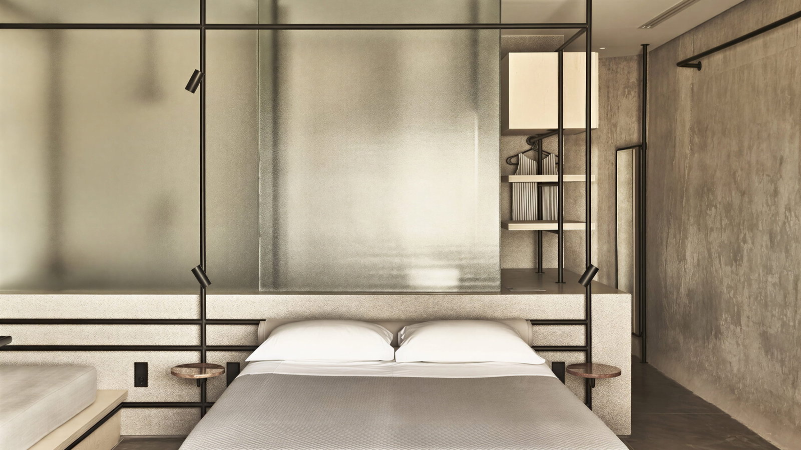 Beachfront WineTank Suite bedroom at Dexamenes Seaside Hotel with a gray bed, frosted glass partition, and beige walls. Shelving unit with books and subtle lighting. Calm ambiance.