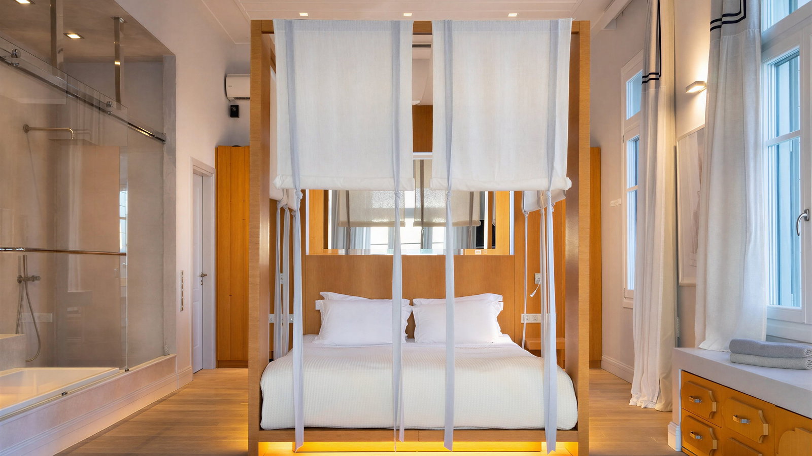 Beachfront villa bedroom with a four-poster bed, white curtains, wood accents, and soft lighting. Glass shower visible; minimal and serene ambiance.