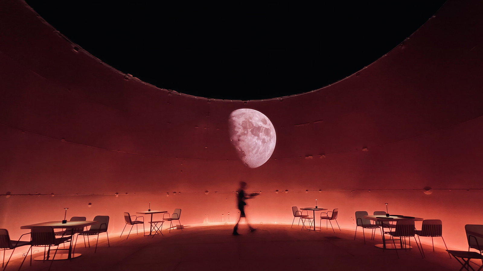 Silo dining room at Dexamenes Seaside Hotel. Silhouette of a person walking in a dimly lit, circular red room with tables and chairs. A large moon projection on the wall. Mysterious mood.