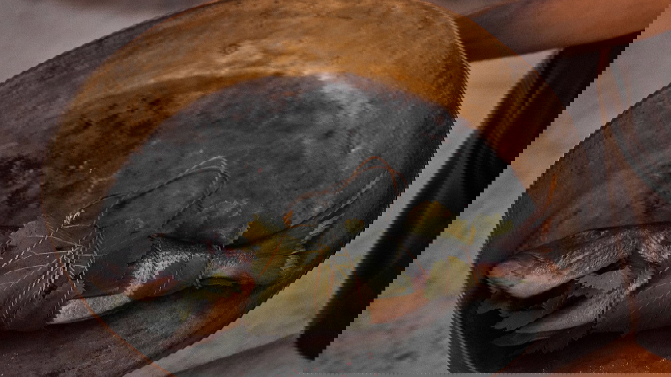 Man holds a whole Branzino at Dexamenes Seaside Hotel in a metal dish with fish wrapped in leaves beside a glowing campfire. Warm tones, evening setting, rustic mood.