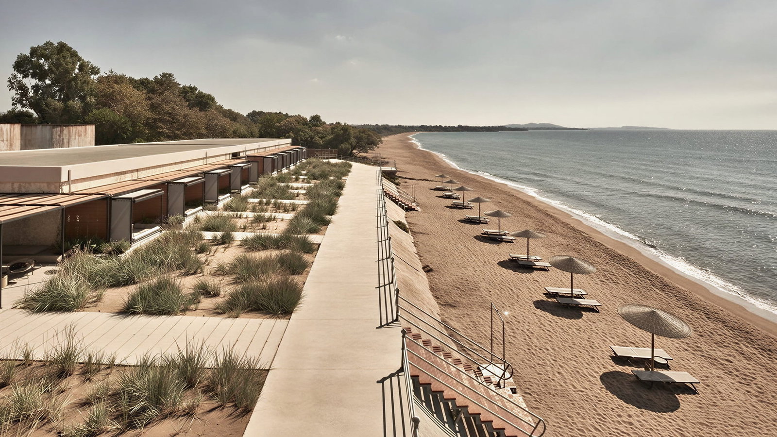 Dexamenes Seaside Hotel beachfront with grass-roofed structures, sandy beach, empty loungers, and parasols under a clear gray sky. Serene atmosphere.