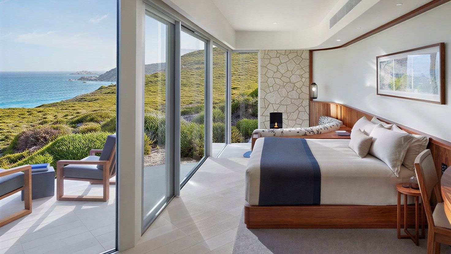 Flinders Suite Bedroom at Southern Ocean Lodge with ocean view, large windows, and modern decor. Features a bed, chaise, chairs outside, and framed art. Calm, sunny ambiance.