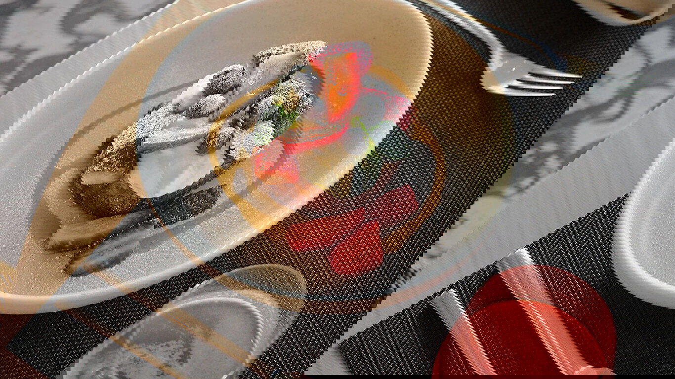 A dessert with strawberries, blueberries, and cream in a white bowl with syrup. Pink drink, cutlery on a black mat. Elegant dining setup.