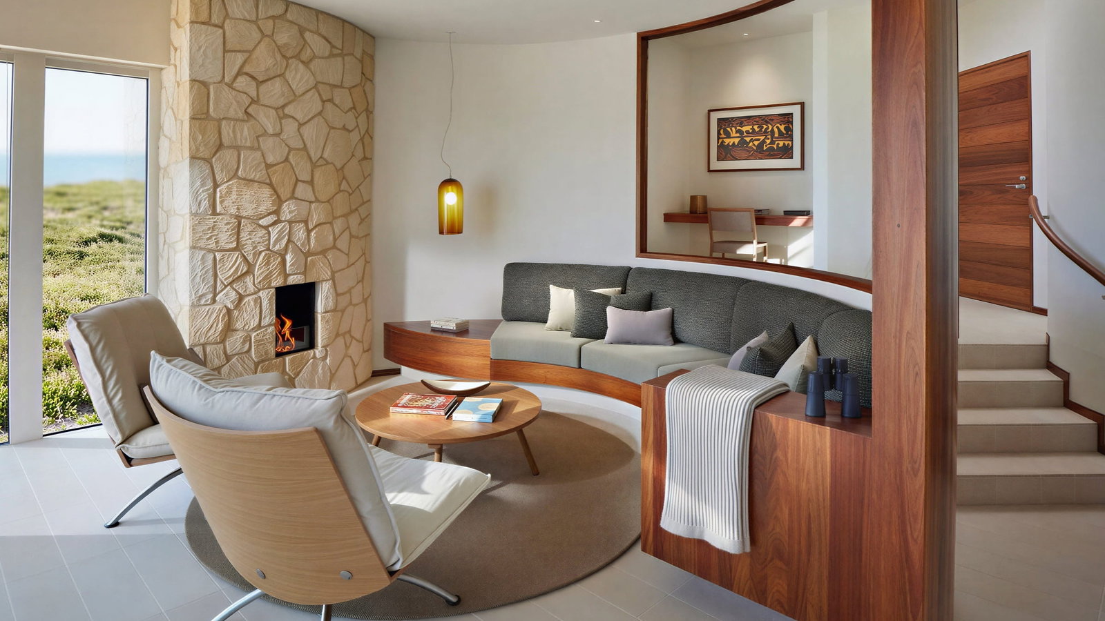 Modern living room of Remarkable Suite at Southern Ocean Lodge with stone fireplace, wooden accents, gray sofa, round table, beige chairs, art on wall, large windows with scenic view.