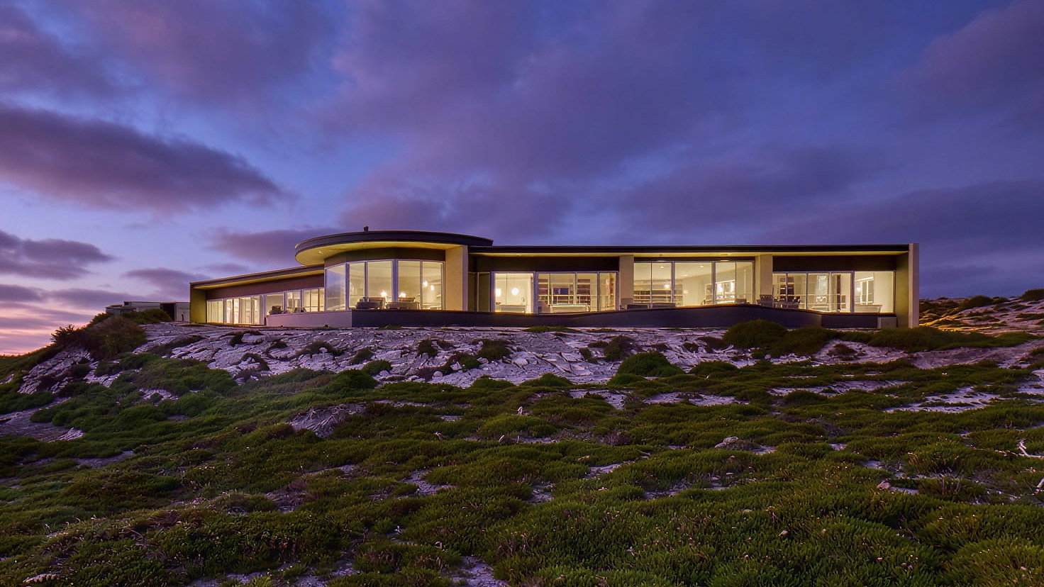 Exterior of Baille Pavilion at Southern Ocean Lodge with large windows, illuminated at dusk. Set on a grassy hill under a purple and pink sky, creating a serene atmosphere.