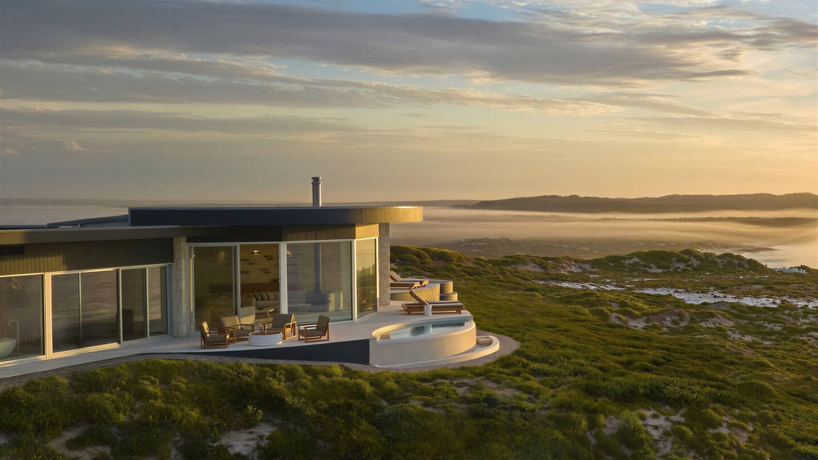 Exterior of Baille Pavilion hot tub at Southern Ocean Lodge with large windows on a green coastal cliff at sunset. Ocean waves in the background, creating a serene mood.