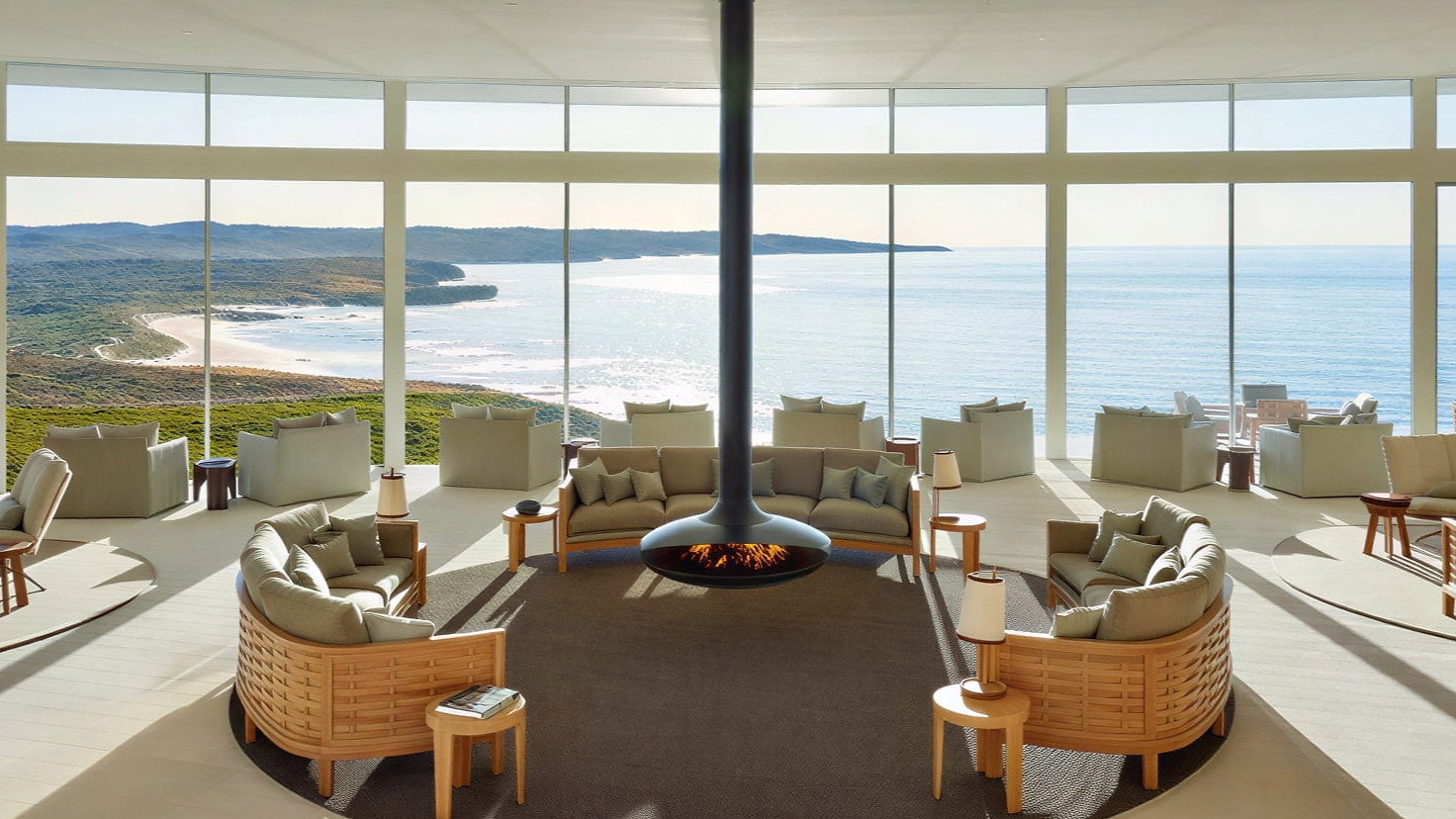 Spacious Great Room with a View at Southern Ocean Lodge with circular seating and central hanging fireplace, overlooking a vast ocean view through large windows. Sunny and serene ambiance.