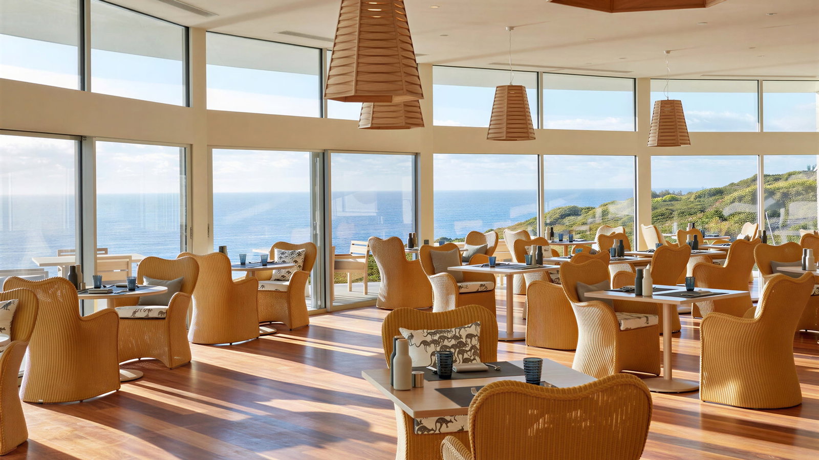 Spacious restaurant dining room at Southern Ocean Lodge with wicker chairs and wooden tables, overlooking ocean view through large windows. Pendant lights add warm ambiance.