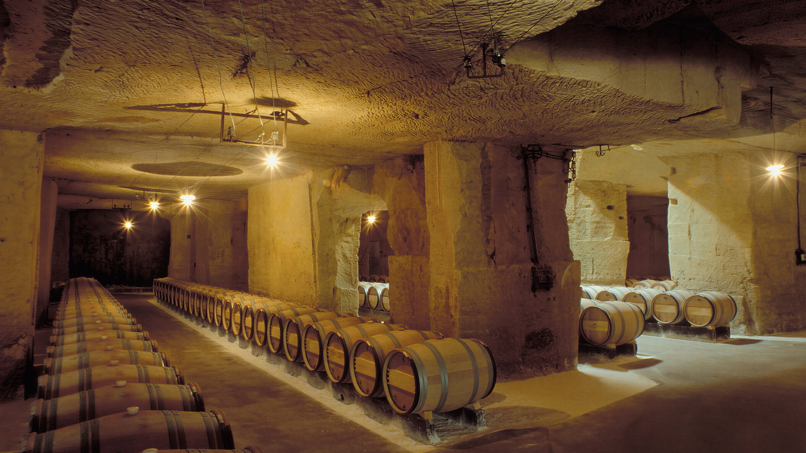 Wine barrels line the dimly lit limestone walls of an underground cellar at Château Ausone. Warm light casts shadows, creating a rustic, tranquil atmosphere.