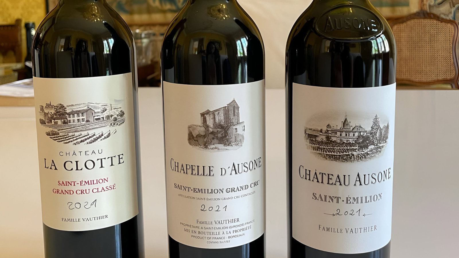 Three wine tasting bottles at Château Ausone labeled Chateau La Clotte, Chapelle d'Ausone, and Chateau Ausone 2021, stand on a table. Tapestry background.