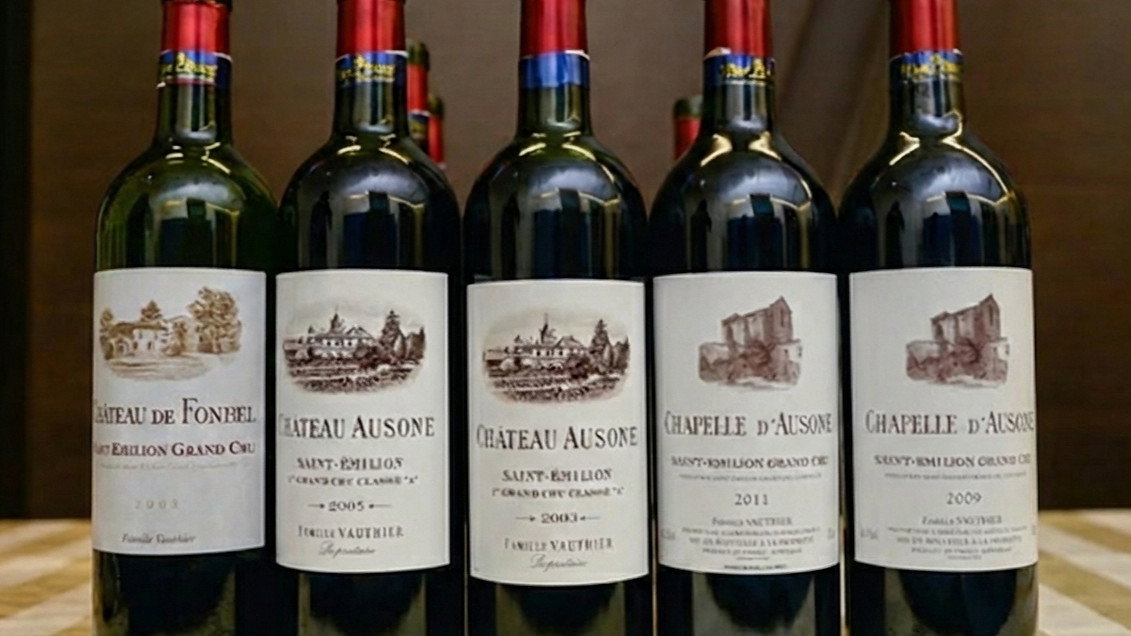 Five Château Ausone wine bottles with red caps on a checkered table; labels read Château Ausone and Chapelle d'Ausone, depicting vintage estates.