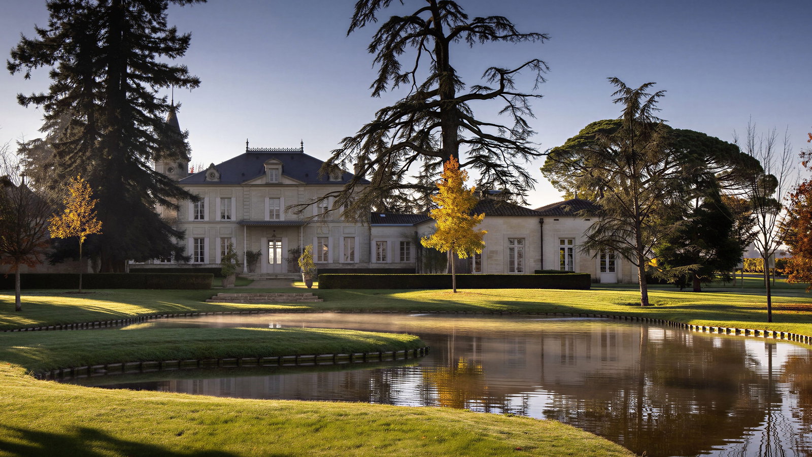 Elegant Château Cheval Blanc estate surrounded by autumn trees, reflected in a calm pond. Sunlight casts shadows on the manicured lawn, creating a serene mood.