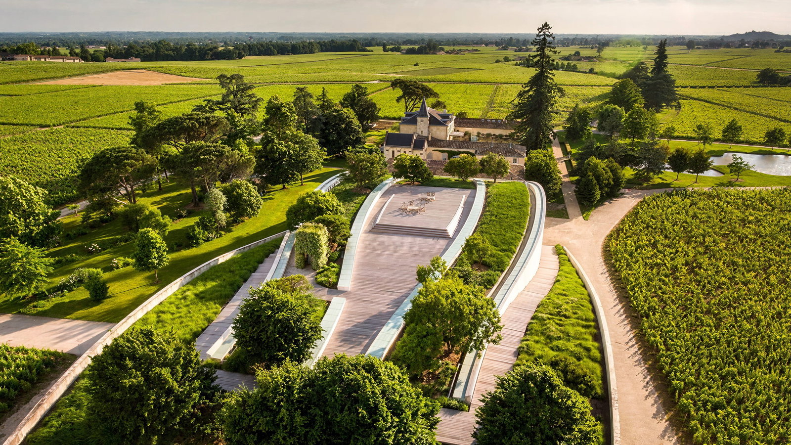 Aerial view of Château Cheval Blanc's modern vineyard estate with lush greenery, a curved wooden terrace, and a scenic horizon under a partly cloudy sky.