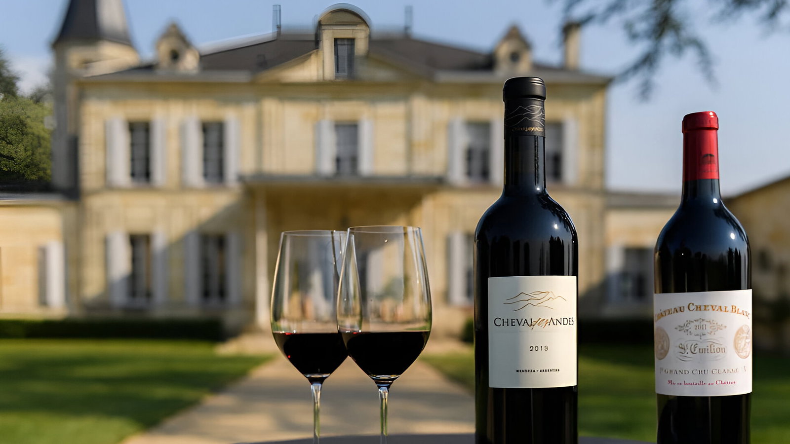 Two wine bottles Cheval des Andes and Château Cheval Blanc Grand Cru and glasses on a table, set in front of a grand chateau with lush greenery. Bright, sunny atmosphere.