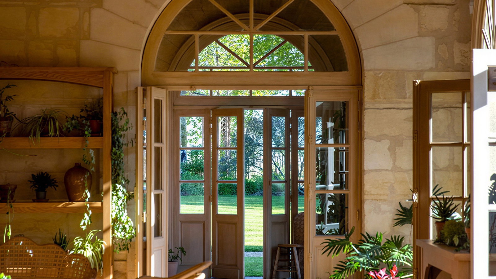 Bright Orangerie reception hall at Château Cheval Blanc with stone walls, arched glass doors, wooden furniture, and potted plants. View of a lush garden outside. Peaceful mood.
