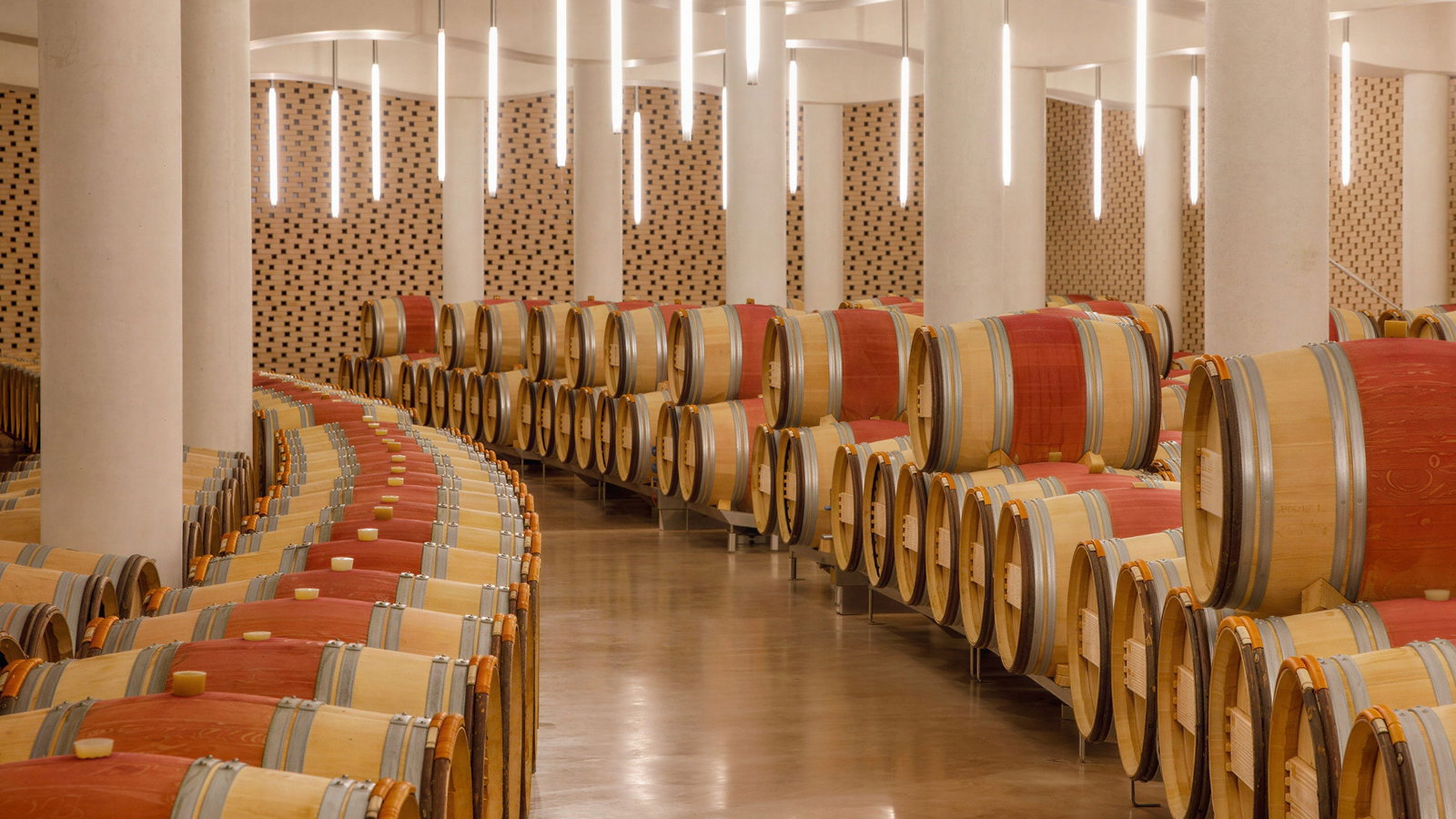 Rows of wooden wine barrels in a modern wine cellar at Château Cheval Blanc with white columns and patterned walls. Bright overhead lights create a sleek ambiance.