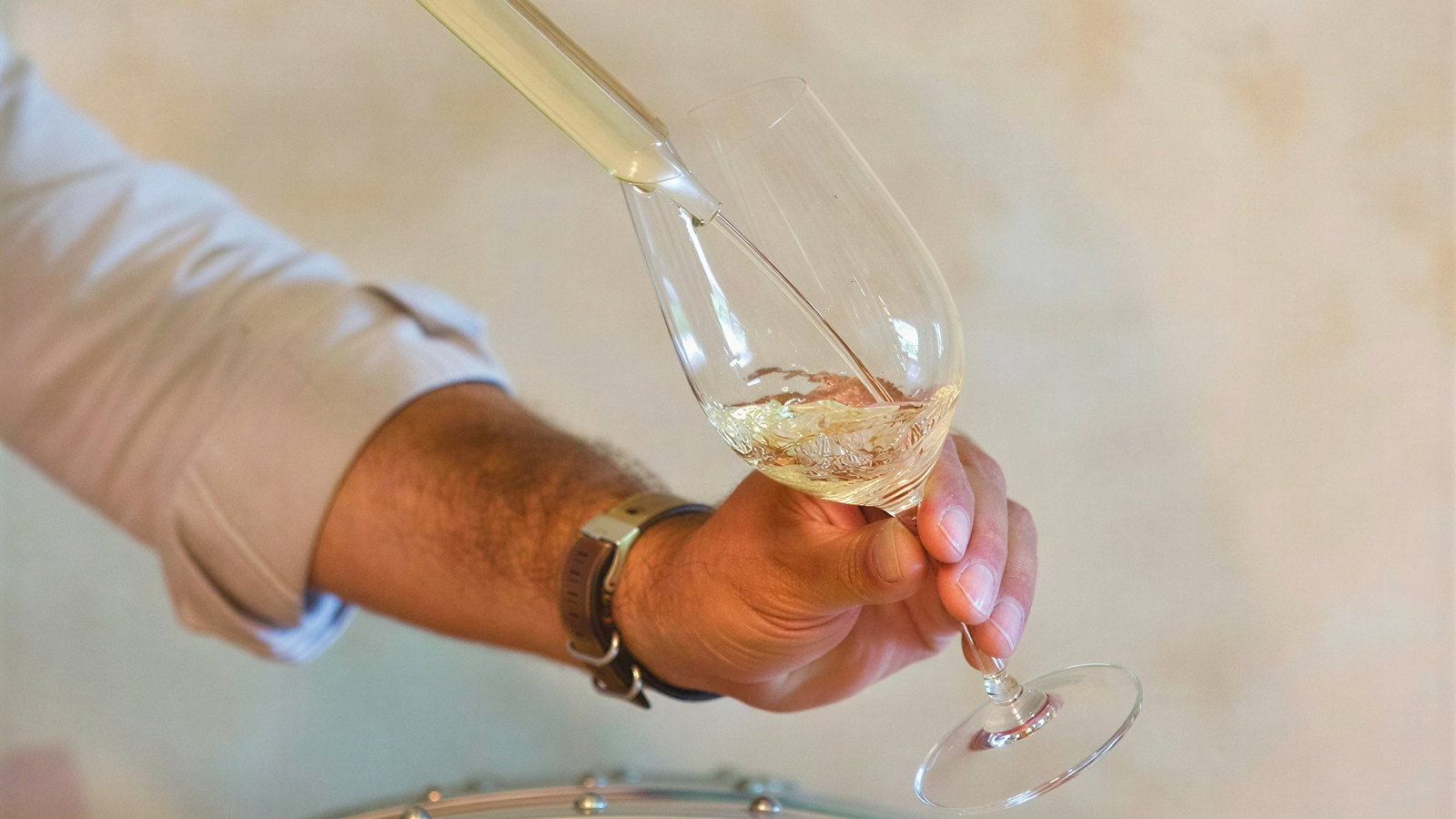 Hand pouring Petite Cheval Blanc wine into a glass from a pipette, above a wooden barrel. The background is a soft beige wall. Mood: calm, focused.