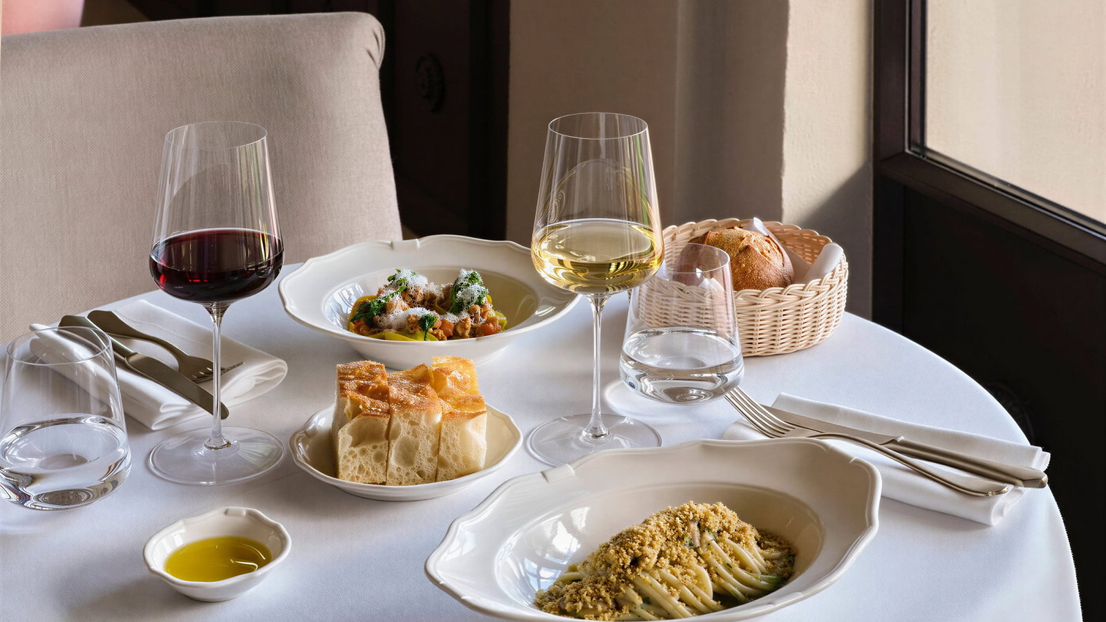 Elegant dinner from La Gamella at Collegio alla Querce with red and white wine, pasta, bread, and stew on a white tablecloth. Warm light from a nearby window.