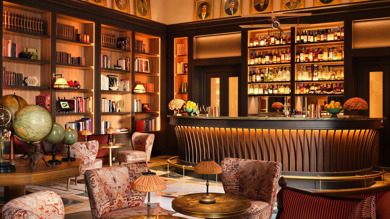 Cozy library Bar Bertelli at Collegio alla Querce with vintage globes, patterned chairs, and warm lighting. Portraits on walls, shelves filled with books, and a stocked bar.