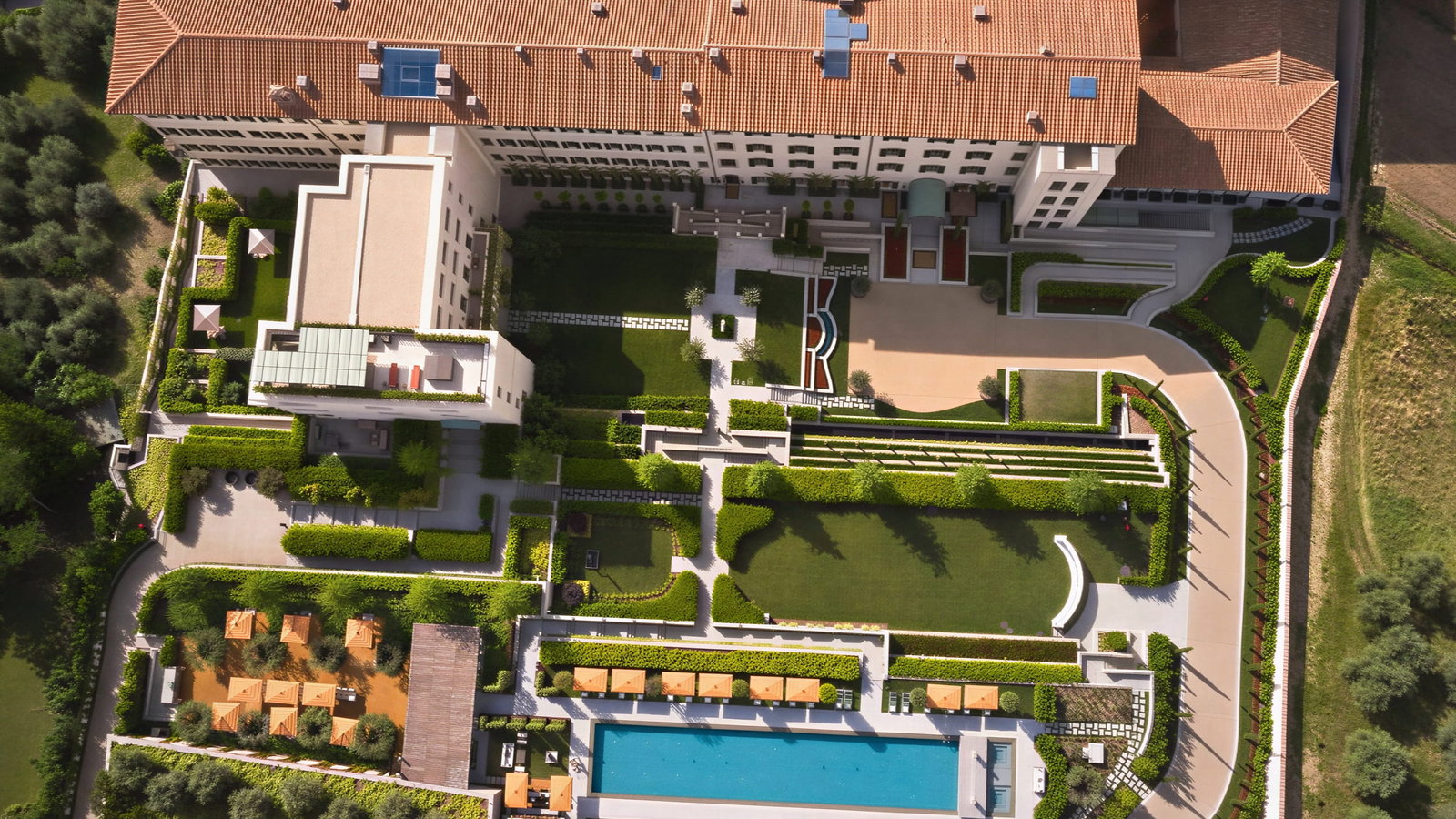Aerial view of pool and gardens at Collegio alla Querce - a large building with terracotta roof, surrounded by lush gardens, a rectangular pool, and sun loungers with orange umbrellas.