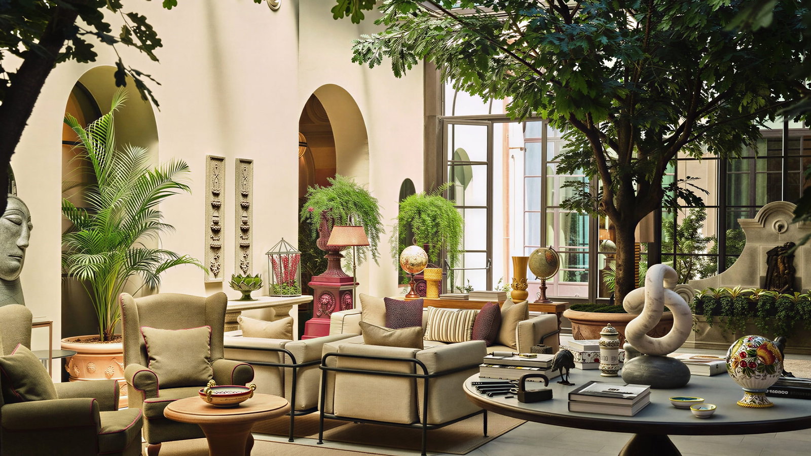 Luxurious indoor Conservatorio Lounge at Collegio alla Querce with plush chairs, round tables, statues, and globes. Large potted plants and windows create a serene atmosphere.