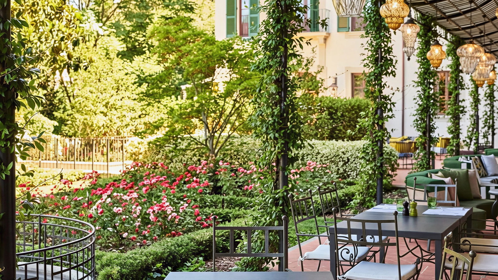 Outdoor patio at Collegio alla Querce with empty black metal tables, surrounded by lush greenery and pink flowers. Overhead lanterns create a relaxed atmosphere.