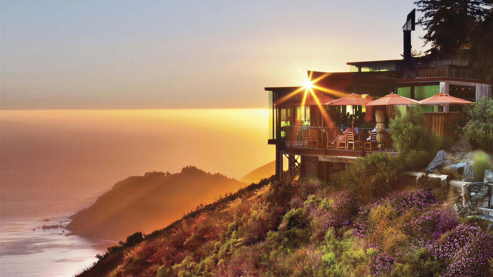 Hilltop restaurant at sunset with diners on a terrace. Ocean and cliffs are visible under a golden sky. Greenery and flowers in the foreground.