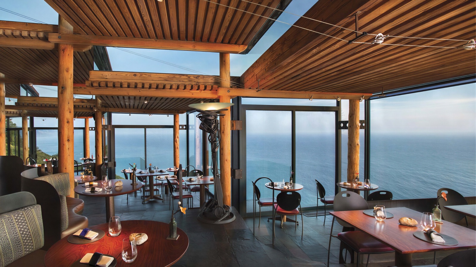 Wooden restaurant interior with ocean view, empty tables set with glasses and napkins. Warm lighting, serene and inviting atmosphere.