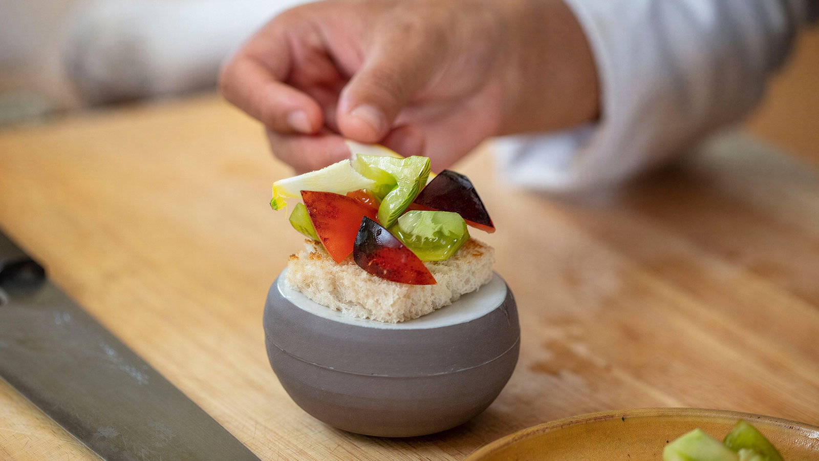 Hand placing colorful salad on bread, atop a round dish on a wooden table. Knife and chopped veggies visible; focused setting.