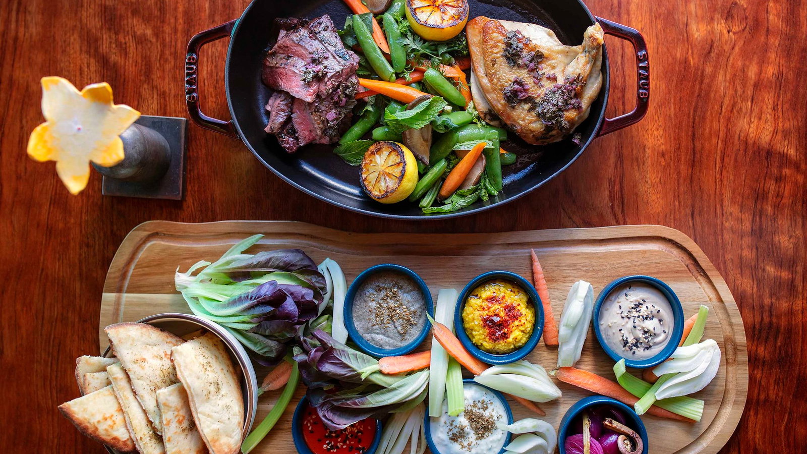 A wooden table with a black dish of grilled meat, veggies, and oranges. A board holds bread, dips, and veggies. Warm tones, cozy setting.