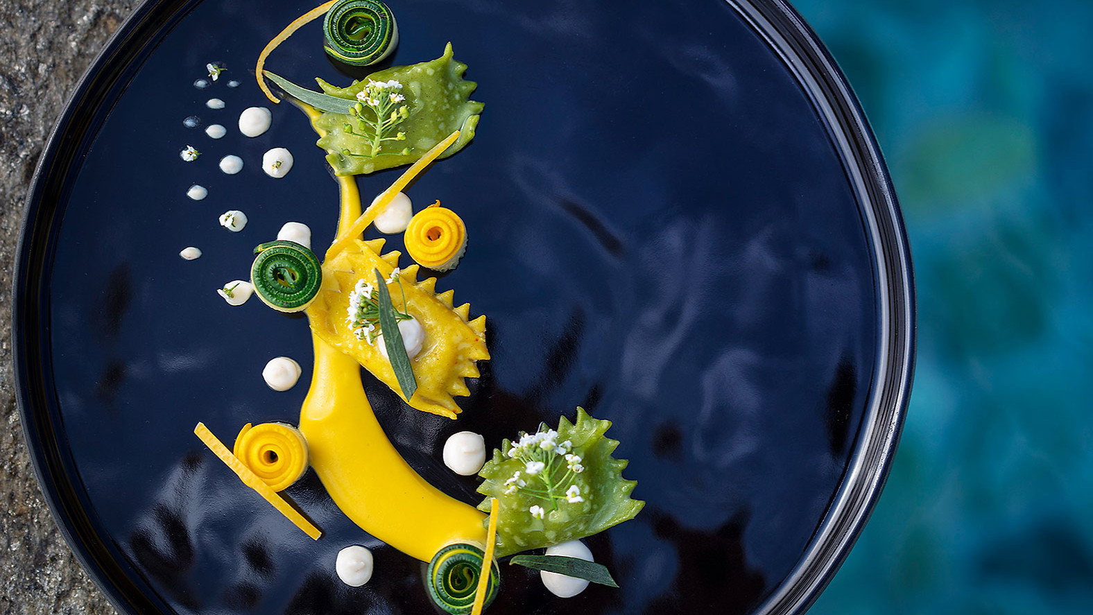 Elegant dish on a black plate with green and yellow pasta, creamy dollops, and garnishes, set against a stone and blue water background.
