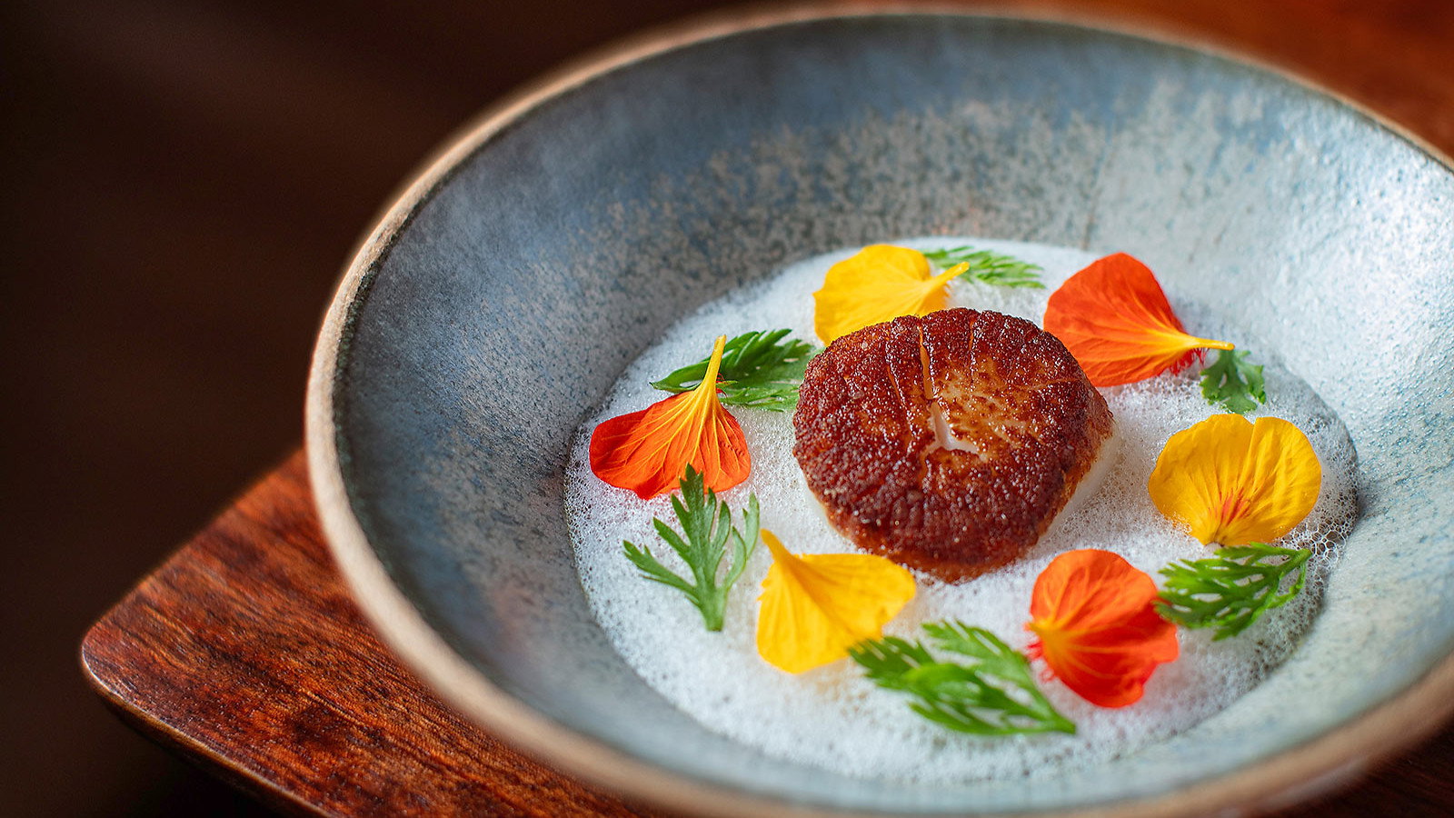 Seared scallop in a gray bowl with foam, garnished with vibrant orange and yellow flowers and green leaves, on a wooden table.