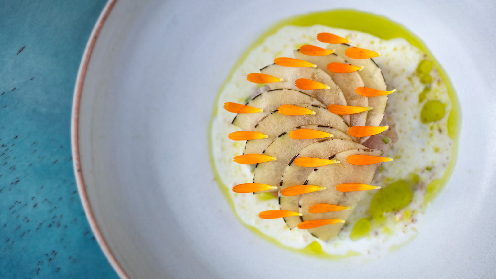 Elegant dish with sliced presentation, topped with orange flower petals on a white plate. Accents of green oil on a textured blue surface.