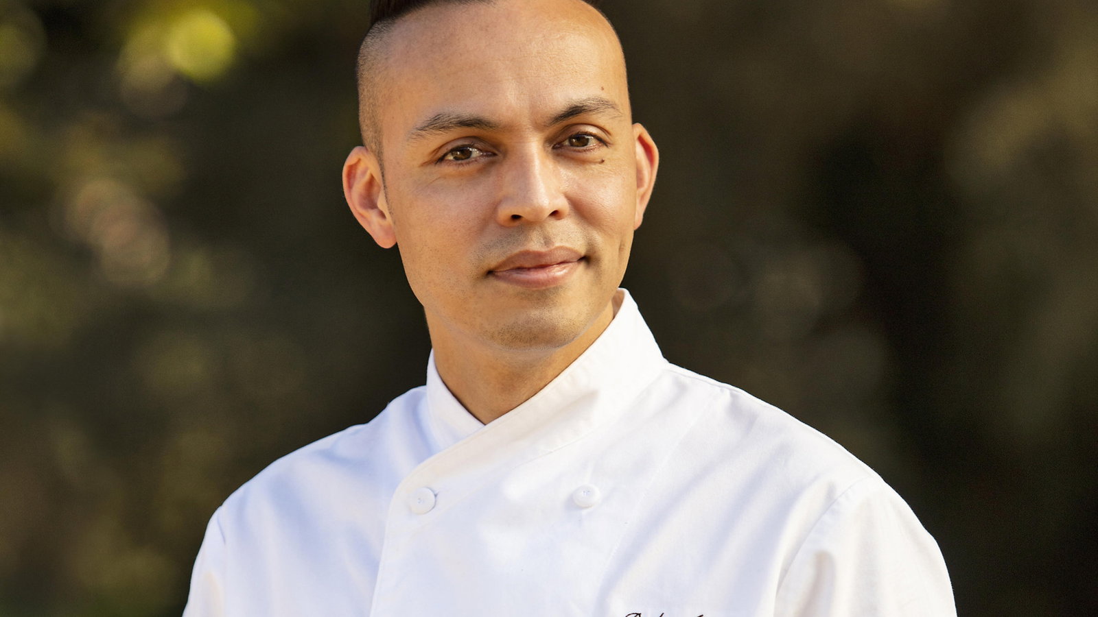 Chef in a white uniform gazes confidently outdoors, with blurred greenery in the background. Name embroidered on the coat.