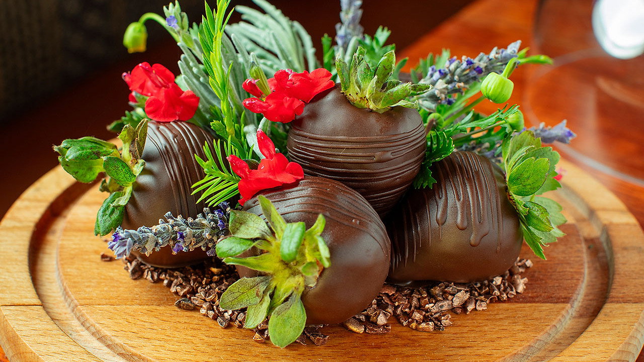 Chocolate strawberries with green leaves and red flowers on a wooden plate, decorated with sprigs of lavender, creating a vibrant display.