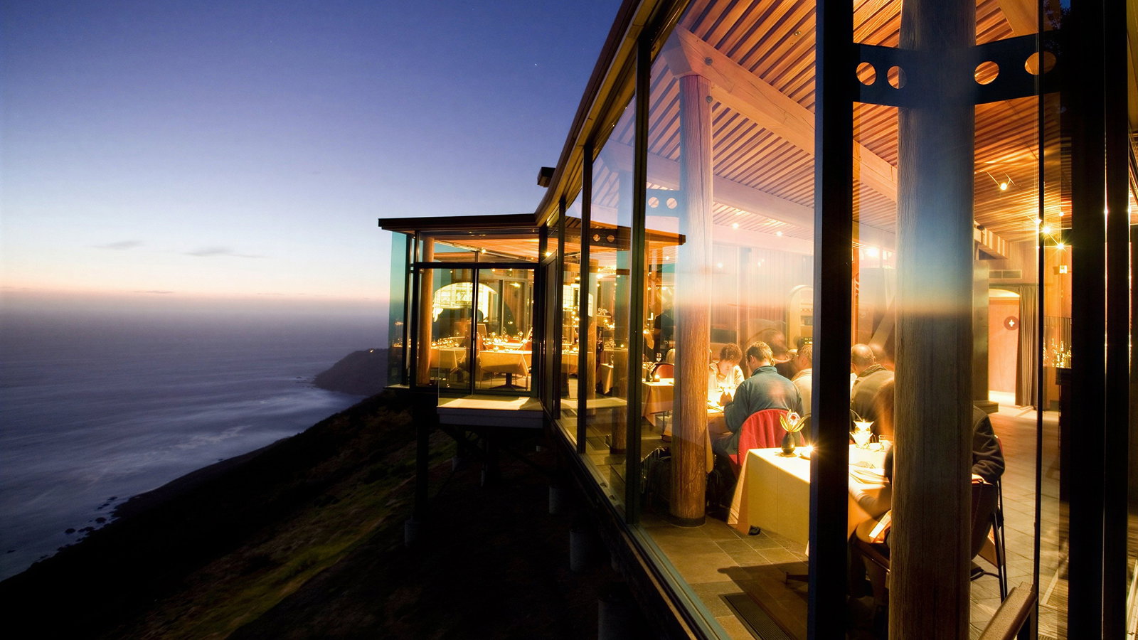 Modern restaurant with large glass windows overlooking a scenic ocean cliff at sunset. Warm interior lighting, guests dining at tables.
