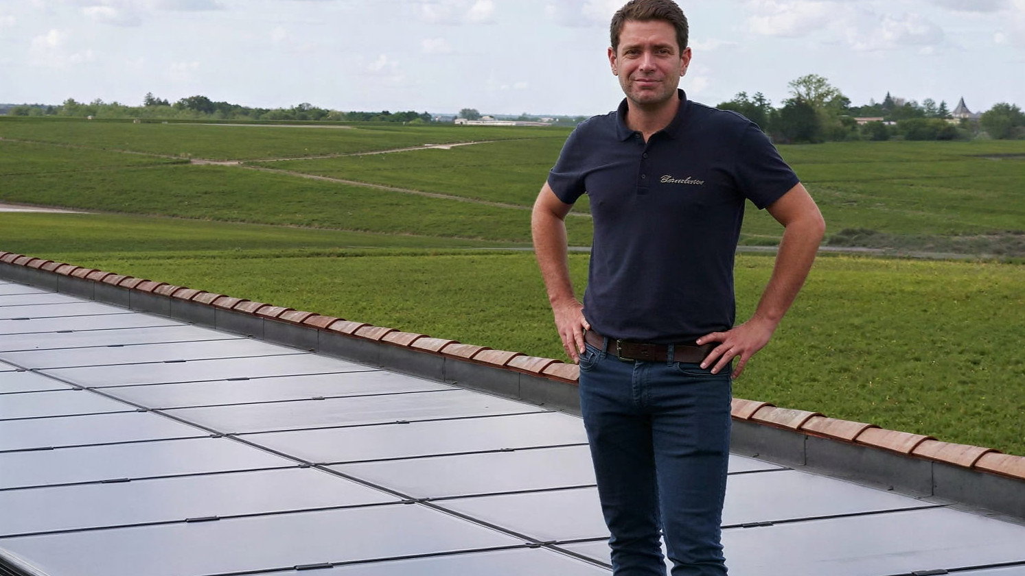 Vincent Decup, winemaker of Château Montrose in blue shirt stands on roof with solar panels, overlooking green fields under a cloudy sky. The shirt reads "Benedicte."
