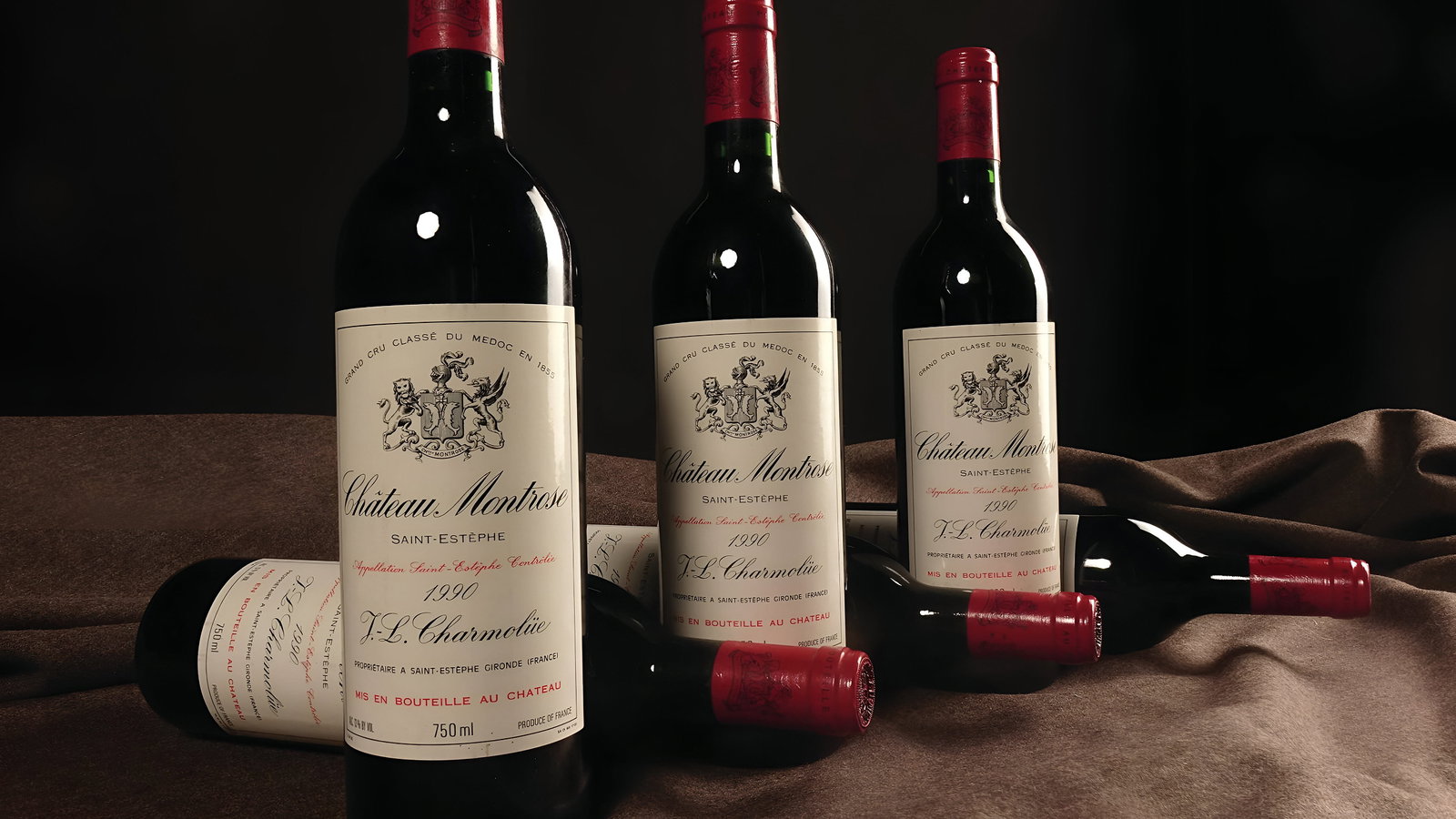 Four 1990 wine bottles with red caps and "Château Montrose" labels on a dark brown cloth. Elegant, dimly lit setting.