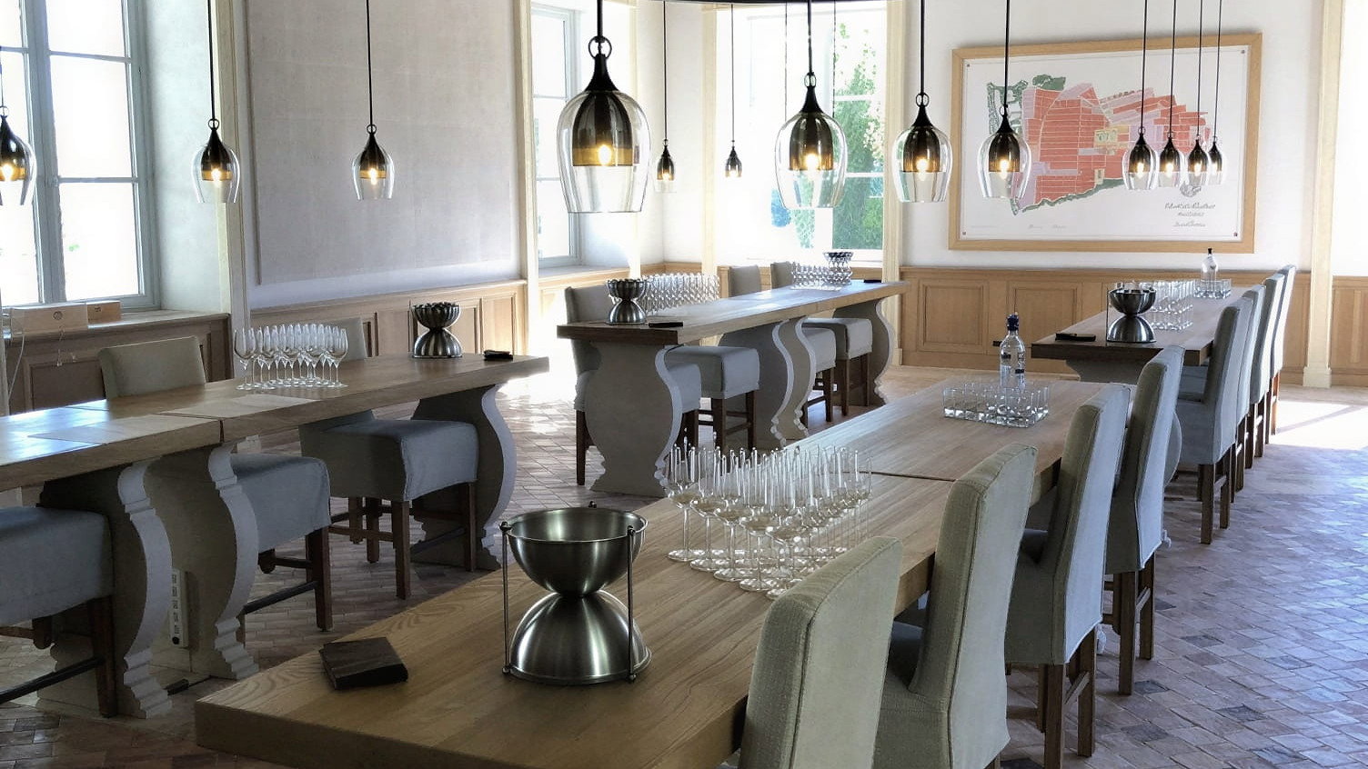 Elegant tasting room at Château Montrose with wooden tables, gray chairs, and hanging pendant lights. Glasses arranged neatly; bright, airy atmosphere.