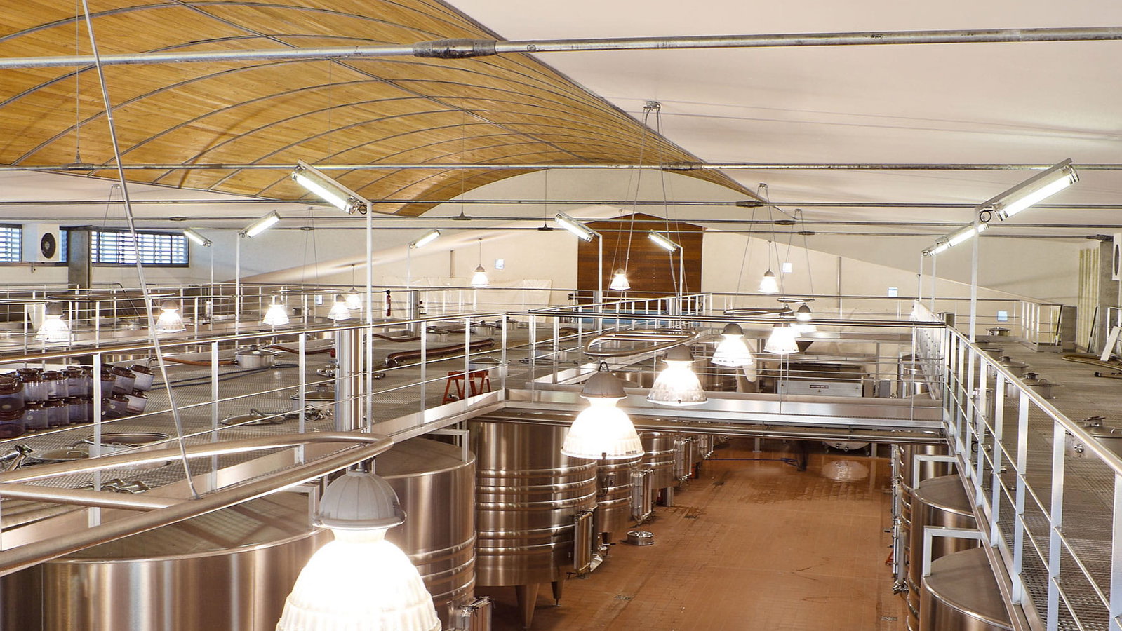 Modern winery vat room interior at Château Montrose with large stainless steel tanks and a wooden-arched ceiling. Bright lighting illuminates the tiled floor. No people present.
