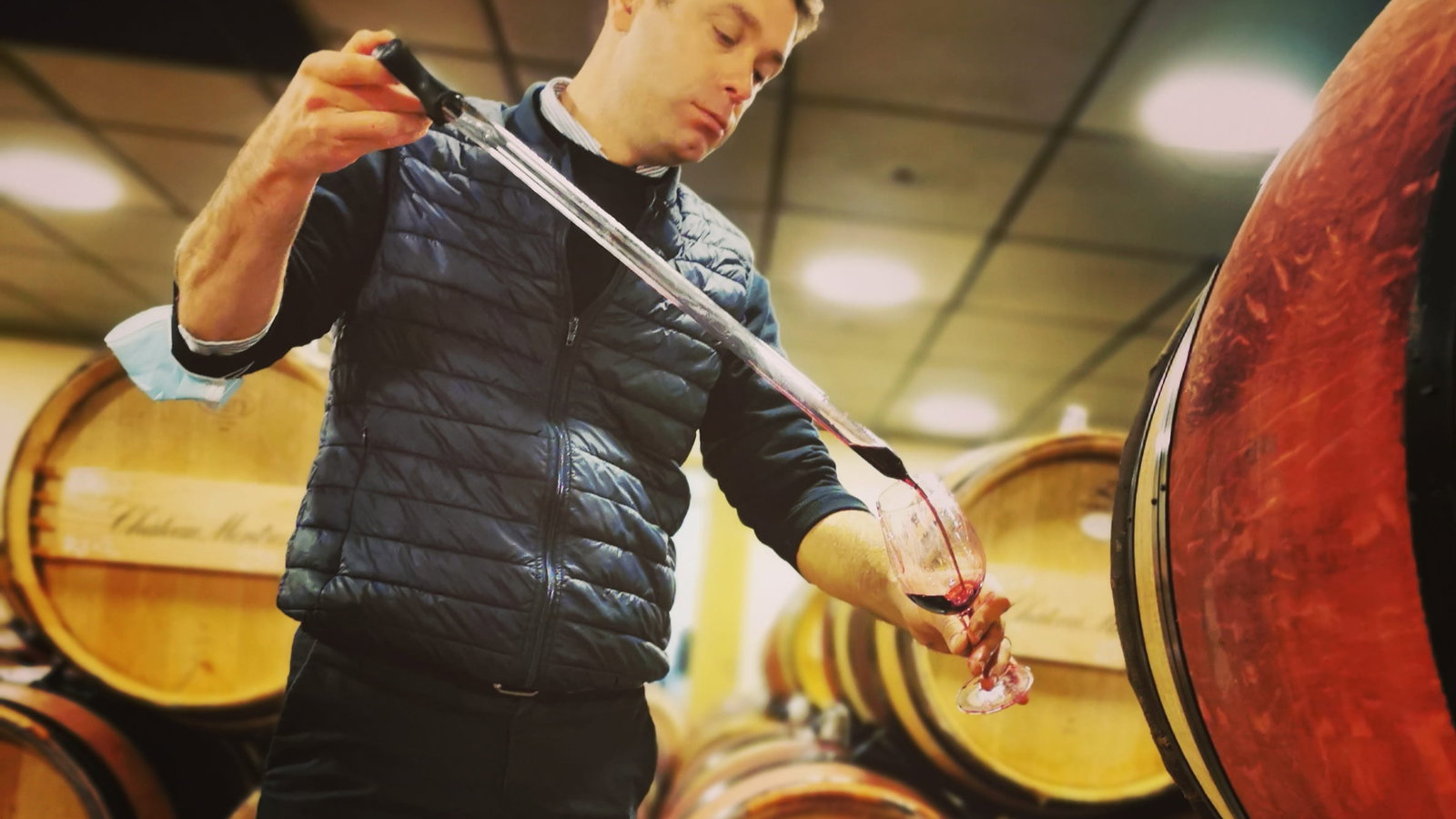 Vincent Decup, winemaker of Château Montrose, in a black jacket tasting wine in a cellar. He uses a wine thief to pour red wine from a barrel into a glass. Wooden barrels in the background.