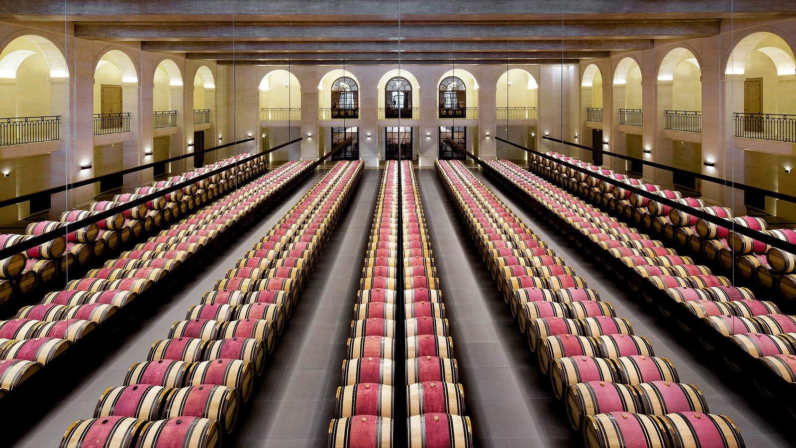 Rows of wooden barrels in the barrel room of Château Montrose with red coverings line a spacious, arched winery cellar. Elegant symmetry, warm lighting, and high ceilings.