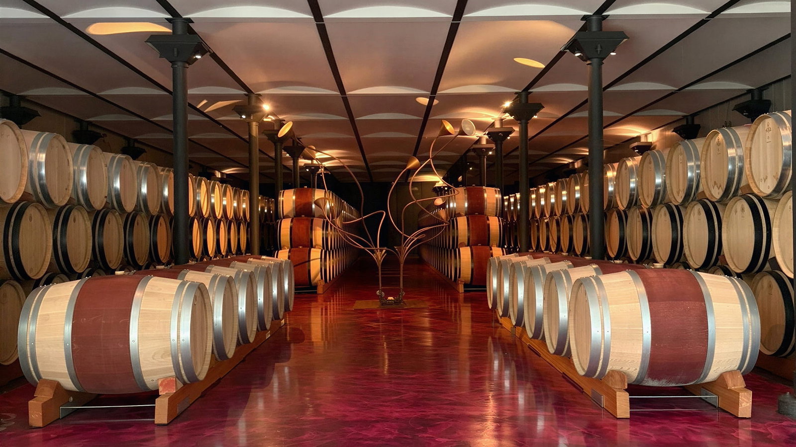 Barrel room at Ornellaia Winery. Wine barrels line a cellar with a reflective red floor and modern ceiling lights, creating a warm and inviting atmosphere.