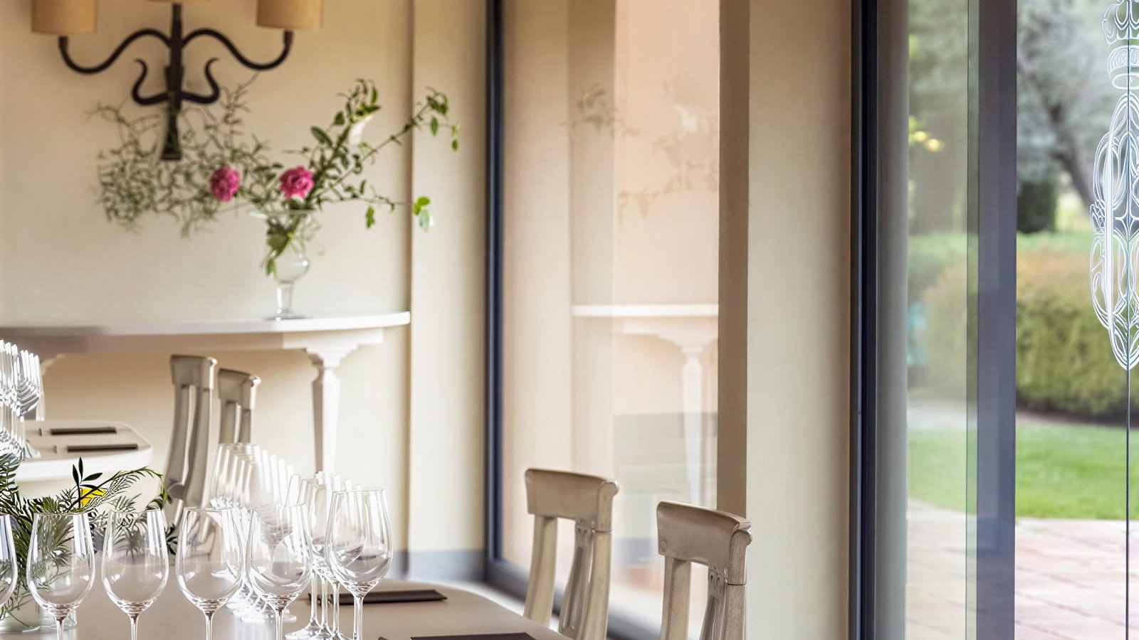Elegant tasting room setup at Ornellaia Winery with rows of empty wine glasses on a table. Vase with pink flowers, beige walls, large window with garden view. Calm ambiance.