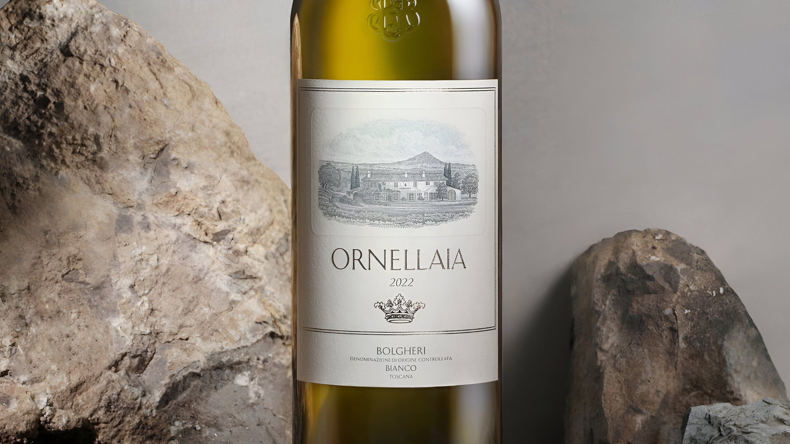 A white wine bottle of Ornellaia 2022 bianco stands on rocks against a gray backdrop. The label features an estate illustration, creating an elegant mood.