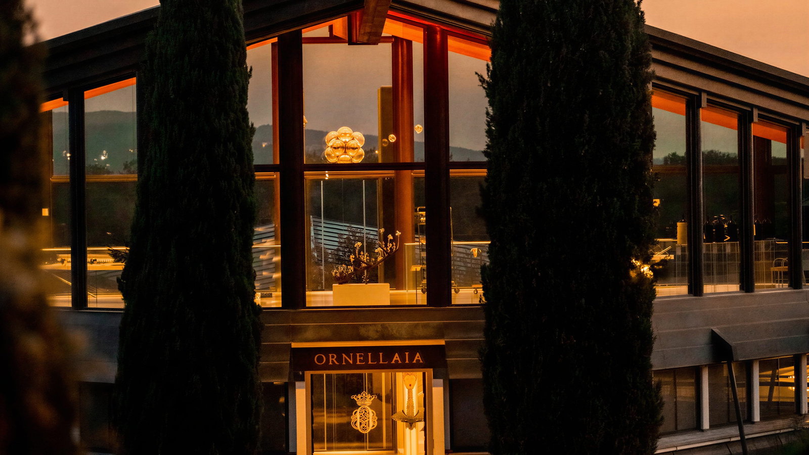 Sunset at Ornellaia Winery's modern glass building at dusk with illuminated entrance labeled "ORNELLAIA," flanked by tall cypress trees and a sunlit path.