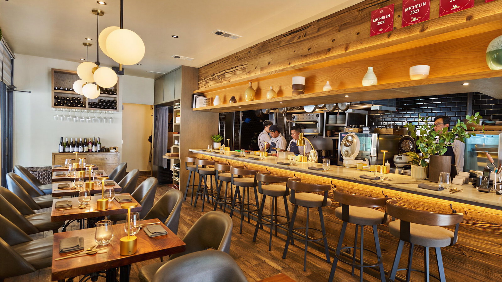 Cozy restaurant with wooden decor, tables set for dining, and chefs working behind the counter. Michelin signs adorn the wall.