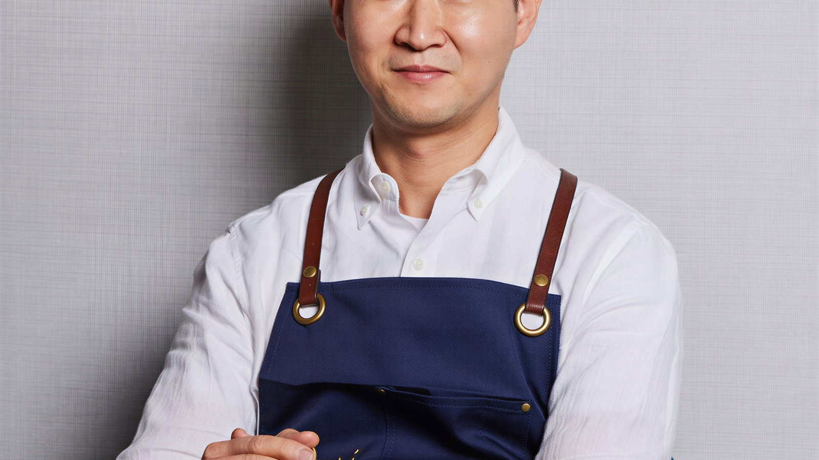 Man in a blue apron sits at a wooden table with folded hands, next to a glass and spoon. Neutral expression. Gray wall and mirror behind.